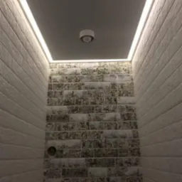 Wnętrze małej łazienki z szarą, podwieszaną konstrukcją sufitową z oświetleniem LED po obwodzie i centralnym punktem świetlnym, ściany wyłożone białą i wzorzystą cegłą.