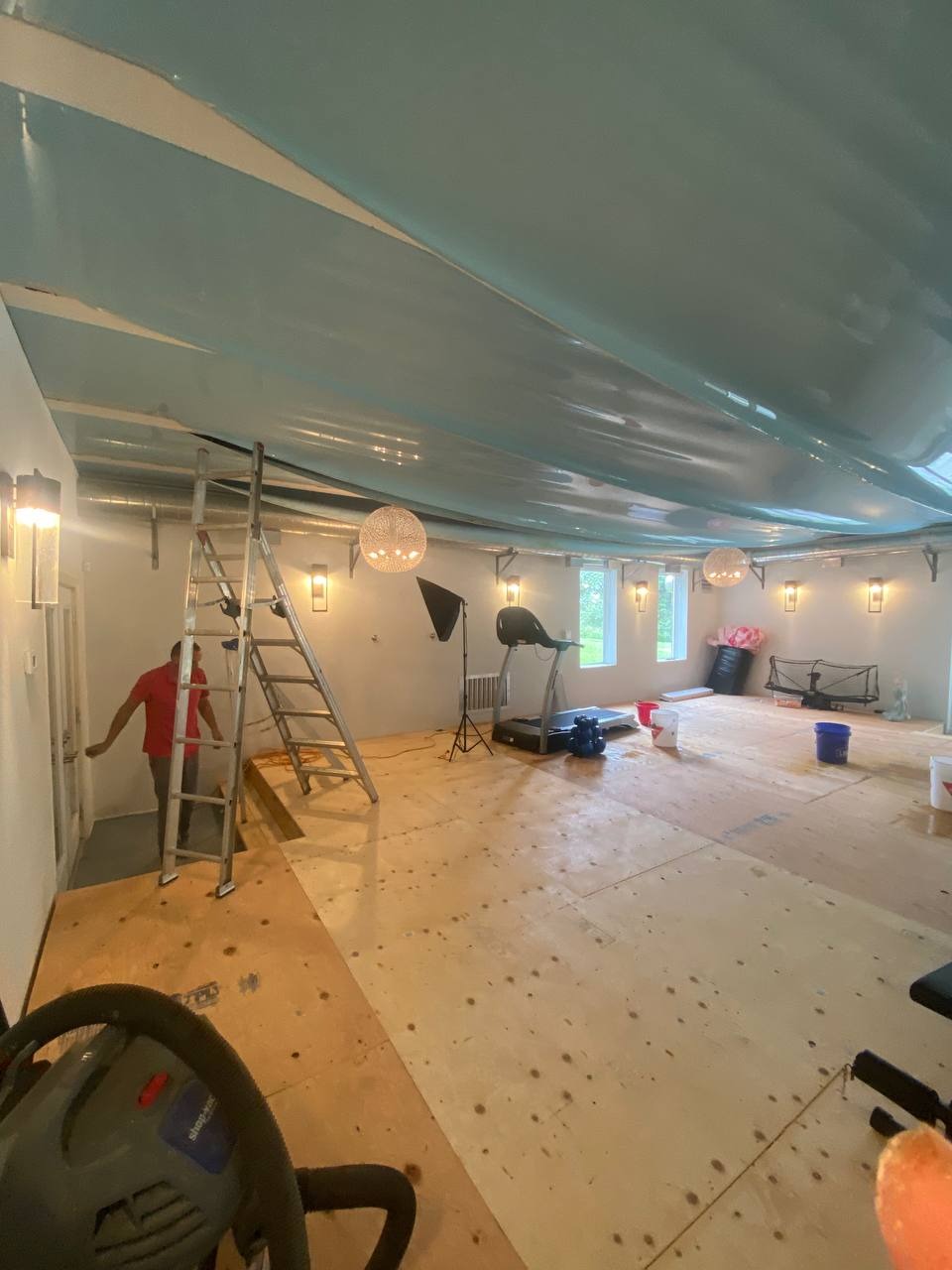 Montaż napinanego sufitu w pomieszczeniu z przygotowaną podłogą, widoczna drabina, lampy kuliste i sprzęt do ćwiczeń.