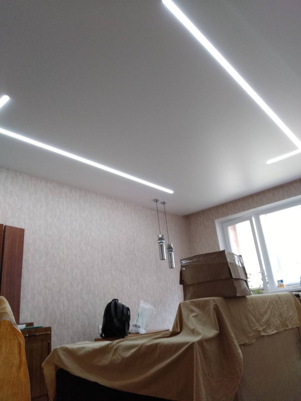 Biały sufit z liniowym oświetleniem LED, widoczne lampy wiszące i meble przykryte materiałem ochronnym.