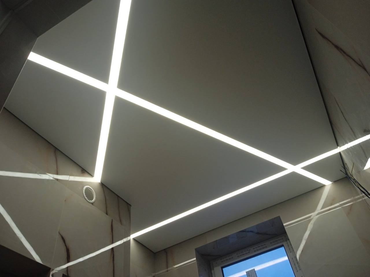 Biały sufit z liniowym oświetleniem LED tworzącym geometryczny wzór, widoczny fragment marmurowej ściany i okno.