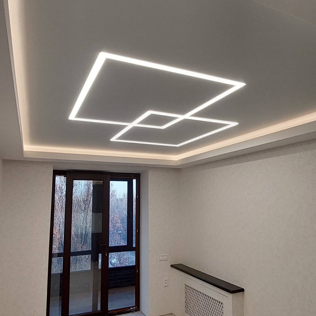 Nowoczesny pokój z białym sufitem, na którym umieszczono geometryczne oświetlenie LED w kształcie dwóch przecinających się kwadratów, widok na przeszklone drzwi balkonowe i grzejnik pod oknem.