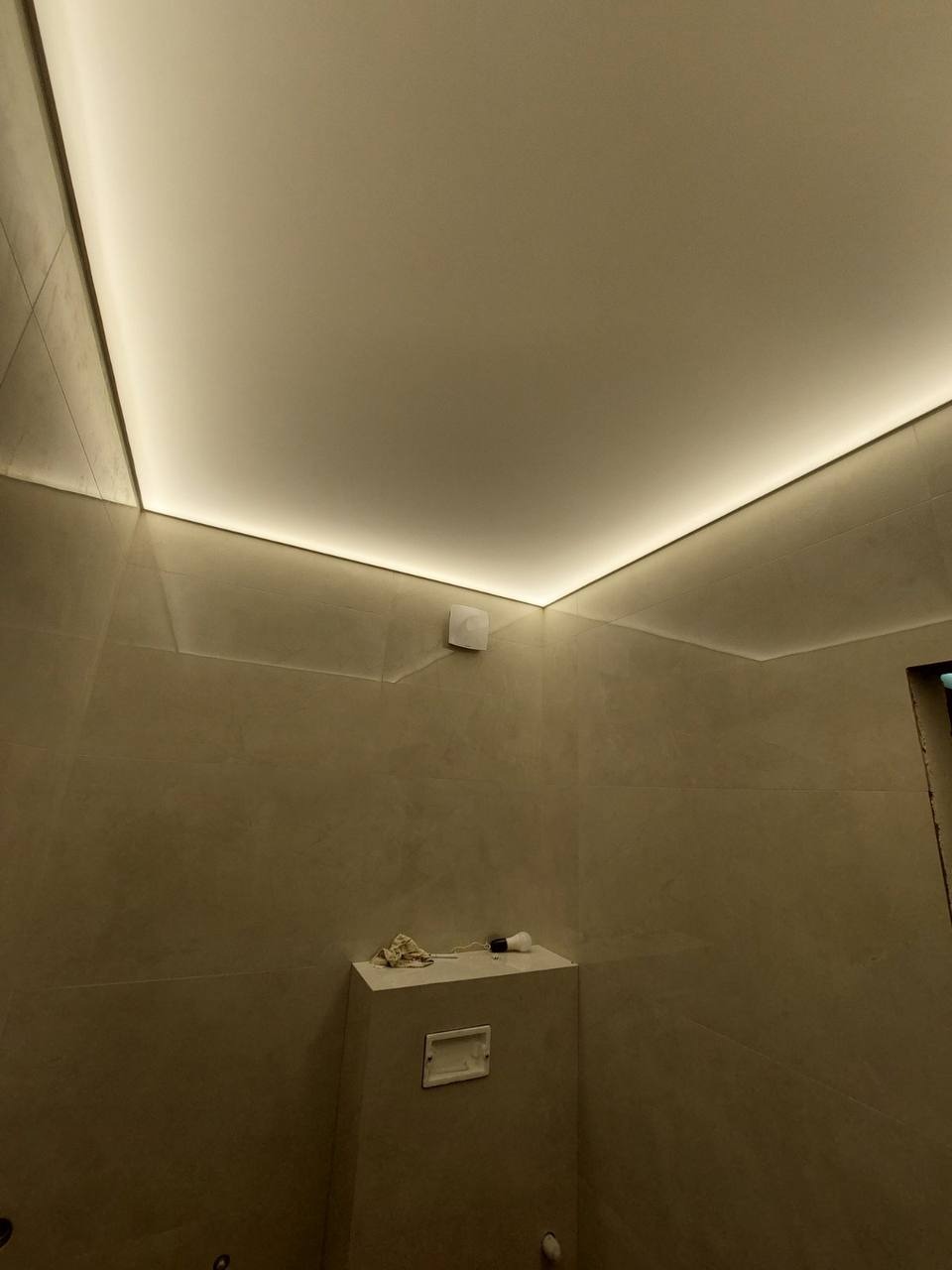 Beżowa łazienka z podświetlanym sufitem napinanym LED, widoczny fragment wentylacji i niedokończone prace wykończeniowe.