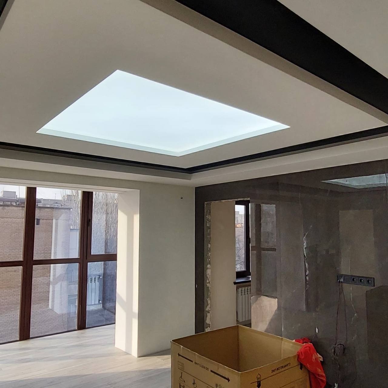 Wnętrze pokoju z kwadratowym, podświetlanym panelem sufitowym, czarnymi listwami dekoracyjnymi i dużym oknem balkonowym.