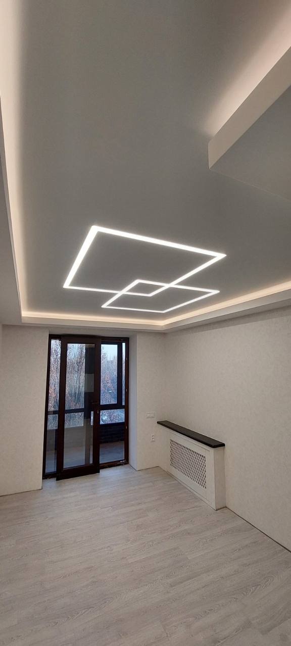 Nowoczesny sufit napinany z geometrycznym wzorem świetlnym, pokój z jasnymi ścianami i drewnianą podłogą, widok na okno balkonowe i grzejnik z półką.