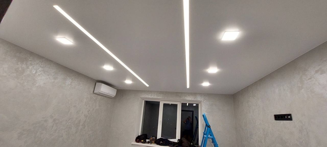 Jasne wnętrze pokoju z szarymi ścianami i białym sufitem z liniowymi i kwadratowymi lampami LED, widoczna drabina i okno.