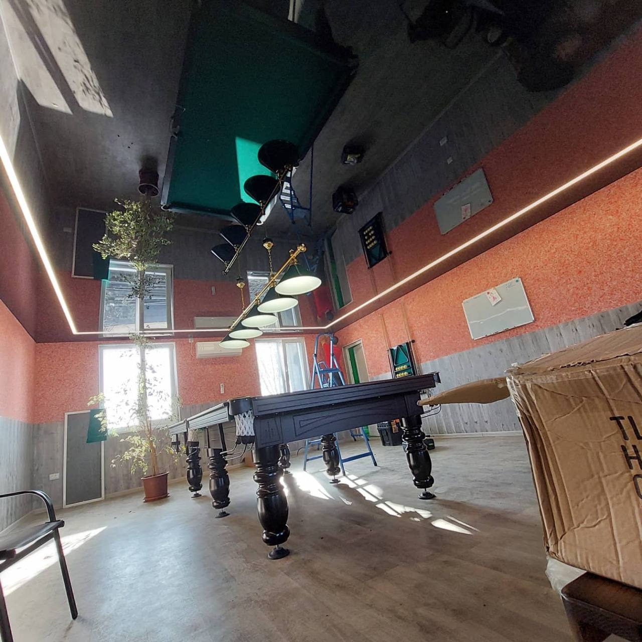 Pokój bilardowy z czarnym stołem bilardowym, oświetlony ledowym paskiem na suficie i ścianach, z widocznym odbiciem stołu bilardowego w błyszczącej powierzchni sufitu.