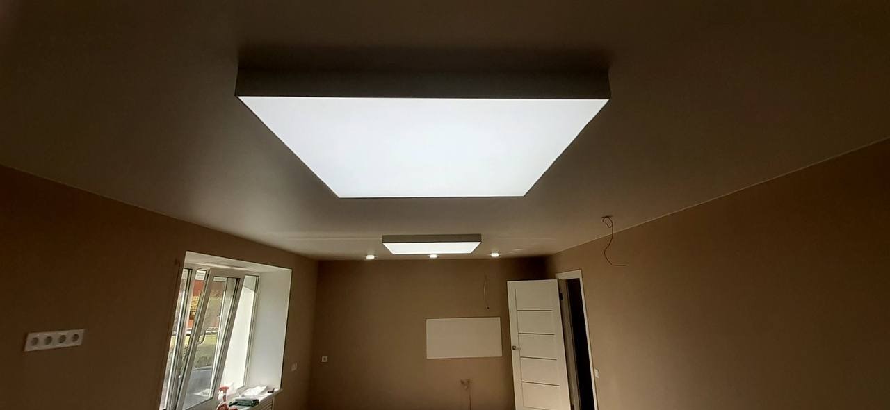 Pokój z beżowymi ścianami i dwoma prostokątnymi, podwieszanymi panelami oświetleniowymi w suficie napinanym, widoczne wystające kable.