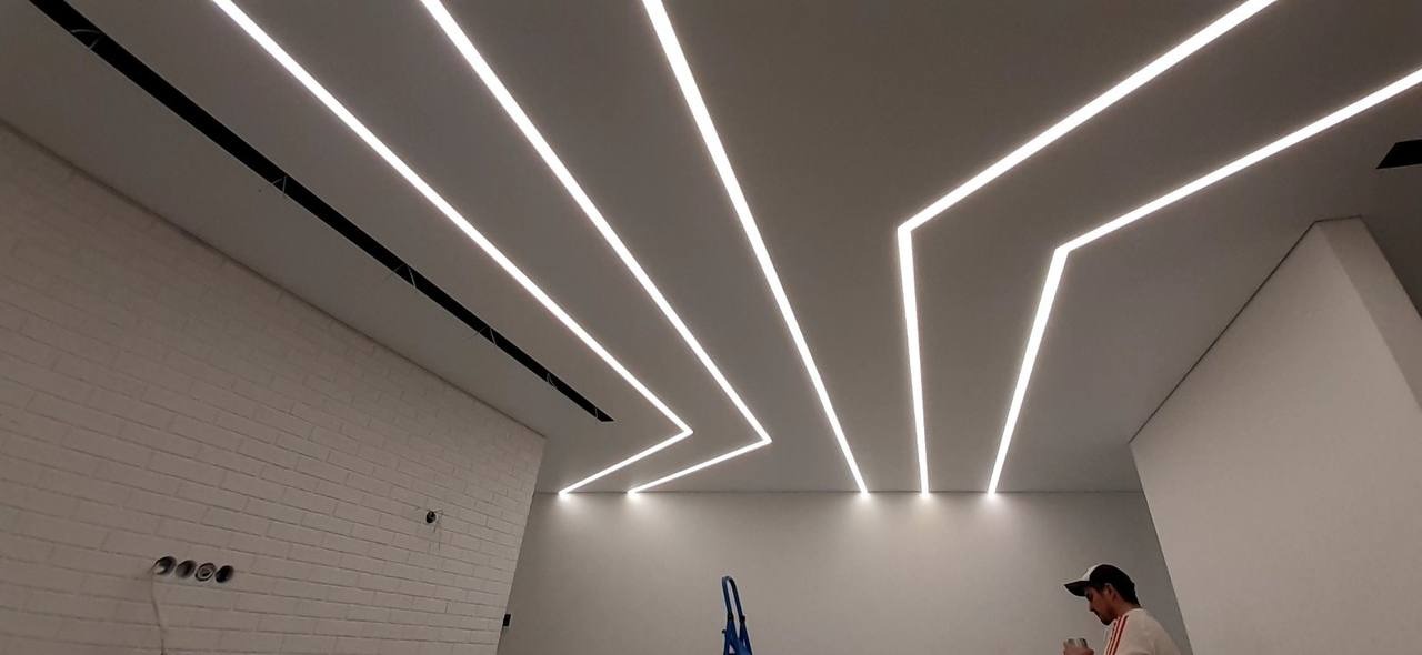 Nowoczesny sufit z liniowym oświetleniem LED tworzącym geometryczny wzór, widoczna ściana z białej cegły i fragment drabiny.