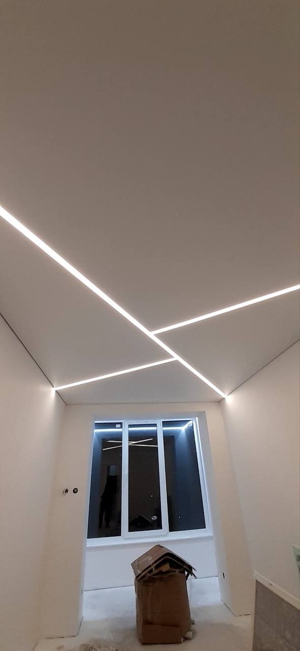 Biały sufit z liniowym oświetleniem LED ułożonym w geometryczny wzór, widok z dołu w surowym wnętrzu z oknem i kartonem na podłodze.