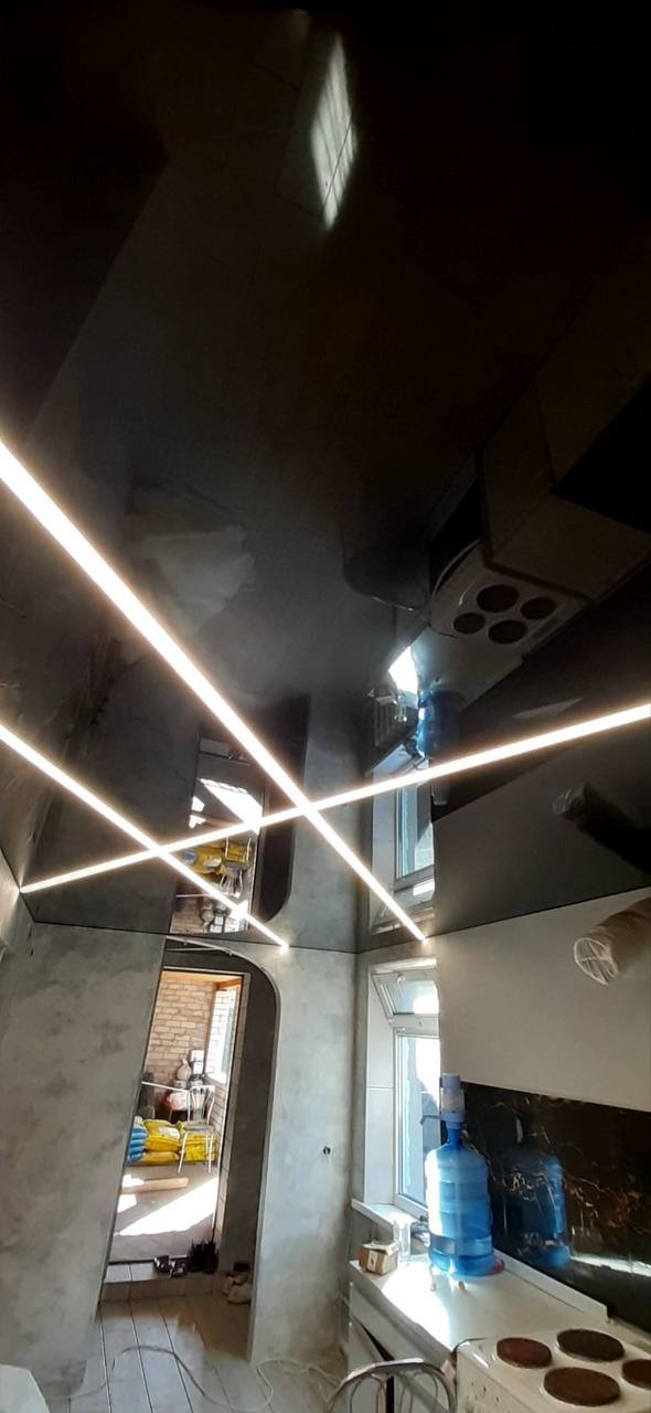 Błyszczący, ciemny sufit napinany z wbudowanymi liniowymi lampami LED krzyżującymi się pod różnymi kątami, widok z dołu w pomieszczeniu z wykończonymi ścianami i widocznym oknem.