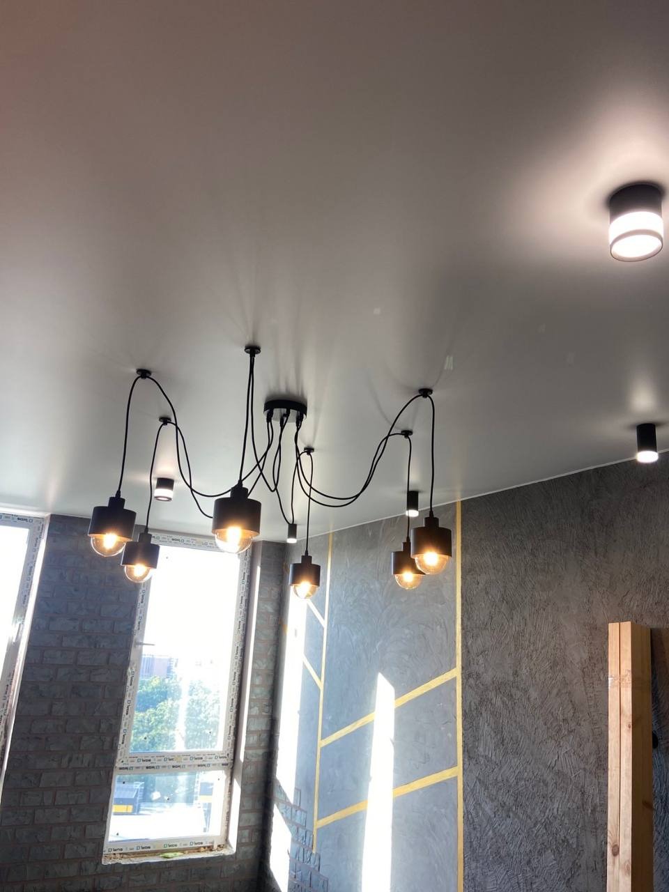 Biały sufit z zamontowanym designerskim, czarnym żyrandolem z wieloma punktami świetlnymi i dodatkowym oświetleniem punktowym, widok na szarą ścianę z geometrycznym wzorem i okno.