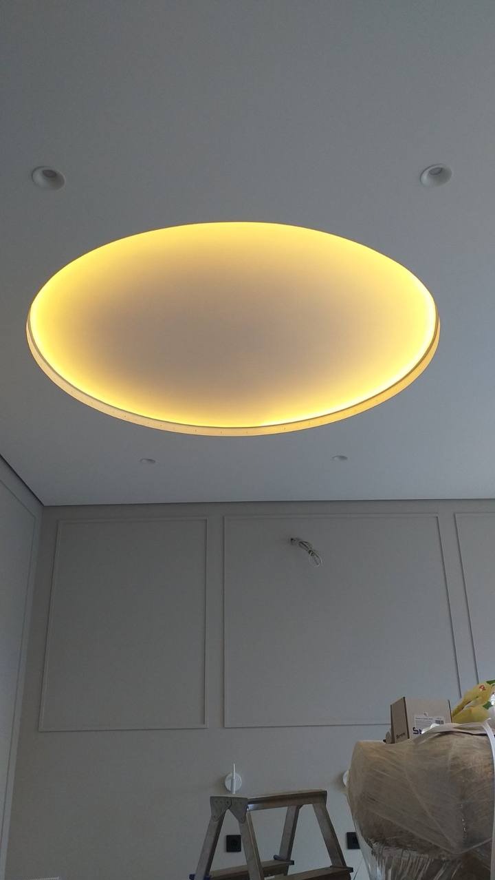 Okrągły panel oświetleniowy w suficie podwieszanym, widoczne małe reflektory punktowe, ściana z dekoracyjnymi panelami w tle, drabina i materiały budowlane w prawym dolnym rogu.