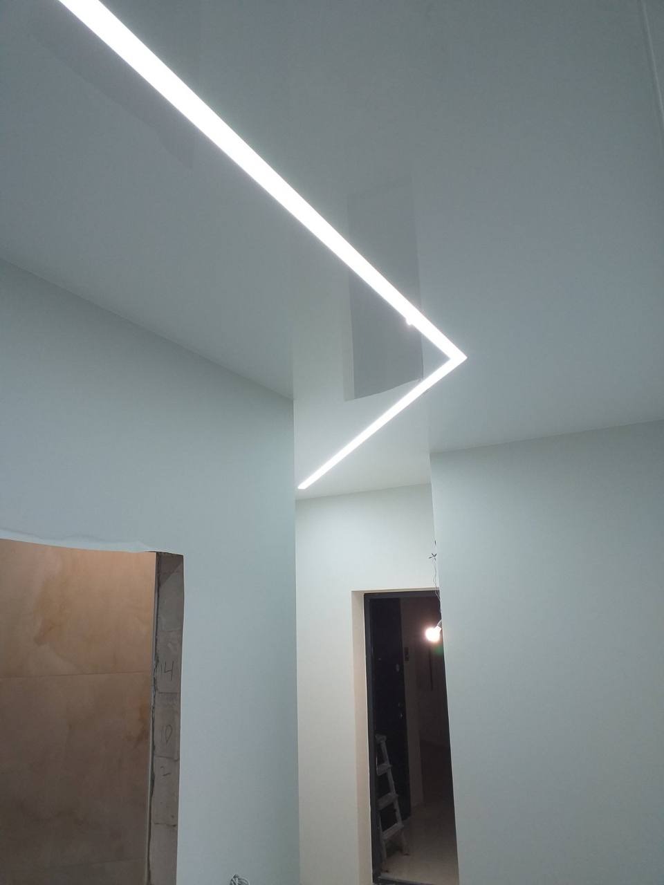 Biały, błyszczący sufit z wbudowanym oświetleniem LED w kształcie litery L, widok z dołu, z widocznymi fragmentami ścian i otworami drzwiowymi.