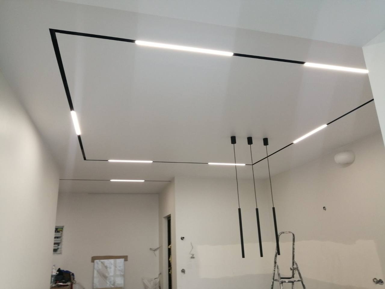 Biały sufit z czarnymi profilami oświetleniowymi LED w kształcie prostokąta, z trzema wiszącymi lampami i drabiną w tle.