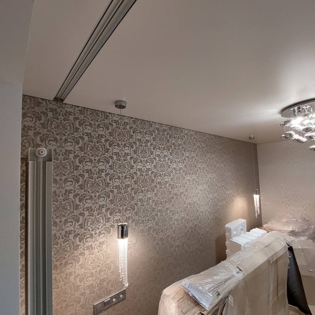 Montaż sufitu napinanego w Warszawie: widoczna szyna montażowa, wzorzysta tapeta na ścianie, nowoczesne oświetlenie, elementy zabezpieczone folią ochronną.
