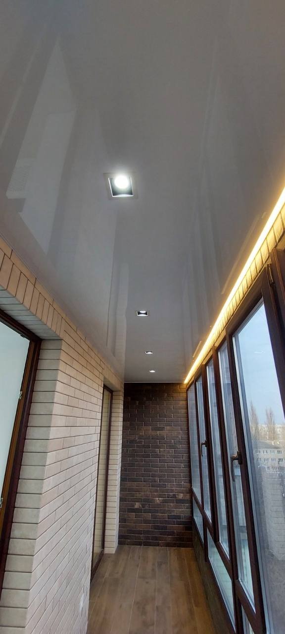 Błyszczący, biały sufit z zamontowanymi lampami punktowymi na balkonie z ceglanymi ścianami i oknami z brązowymi ramami.
