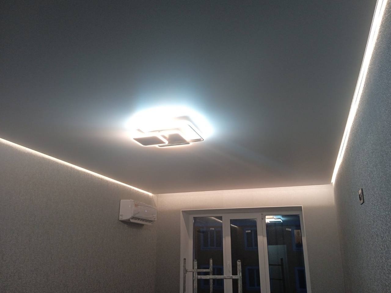 Jasnoszary sufit z oświetleniem LED po obwodzie i nowoczesną lampą sufitową, widoczny klimatyzator i okno balkonowe z drabinką.