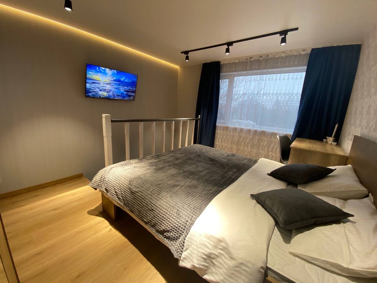 Sypialnia z jasną podłogą, szarą narzutą, ciemnymi zasłonami i telewizorem na ścianie z podświetleniem LED przy suficie.