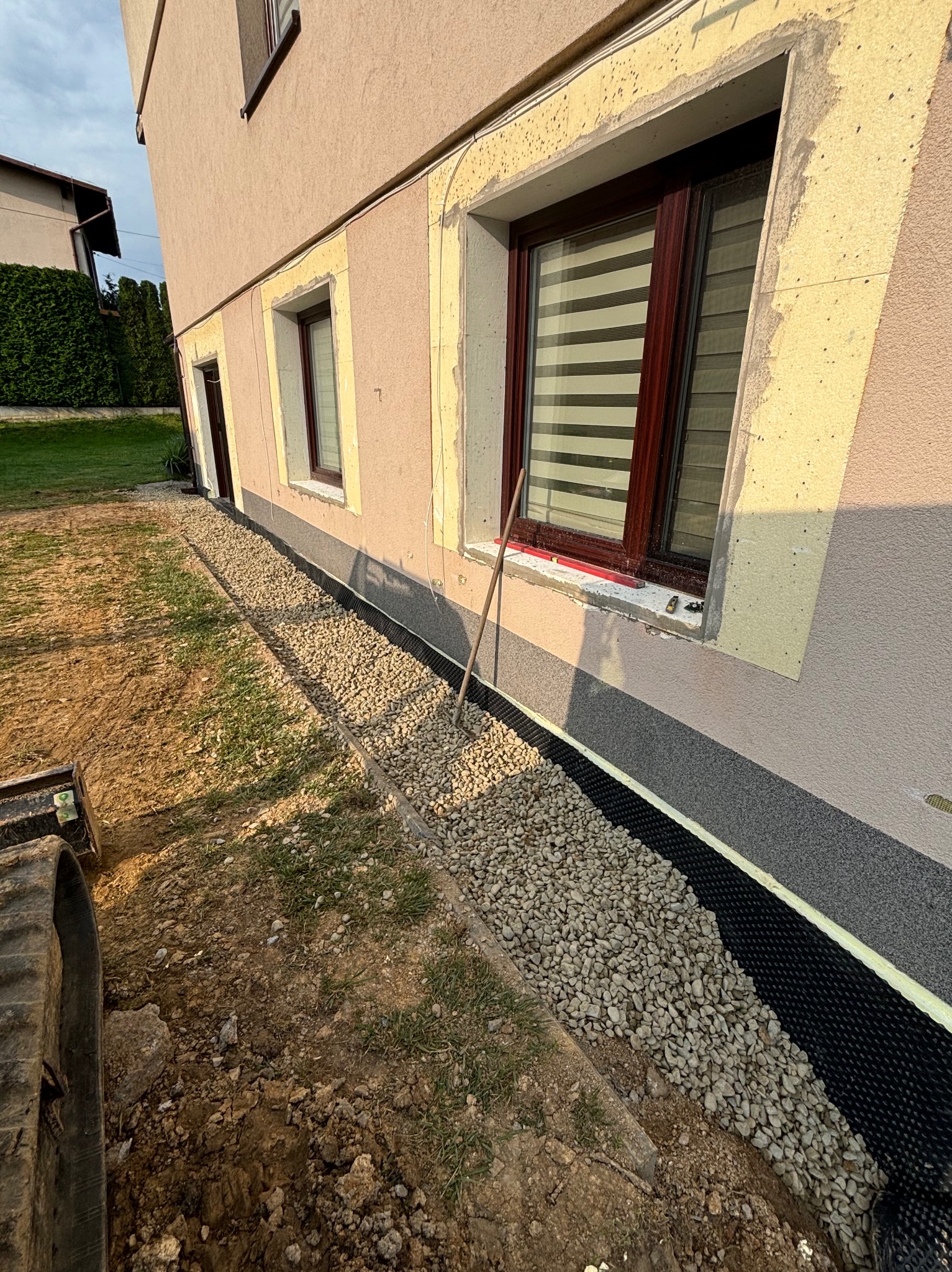 Fundament budynku z ukończonym drenażem: folia kubełkowa, warstwa żwiru, częściowo odkryty wykop, gąsienica koparki w rogu kadru.