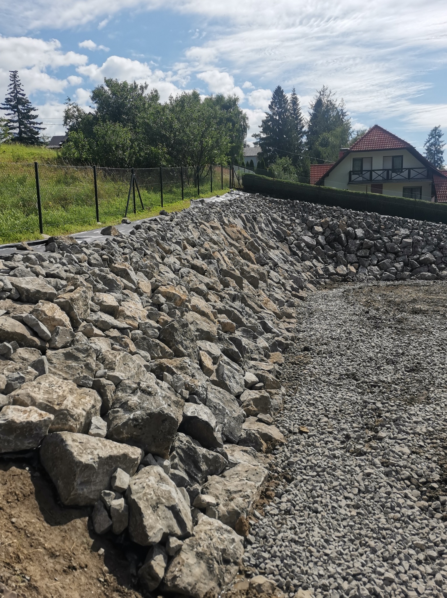 Skarpa wzmocniona dużymi kamieniami i drobnym żwirem, z widoczną czarną folią izolacyjną u góry, w tle dom z czerwonym dachem i drzewa.
