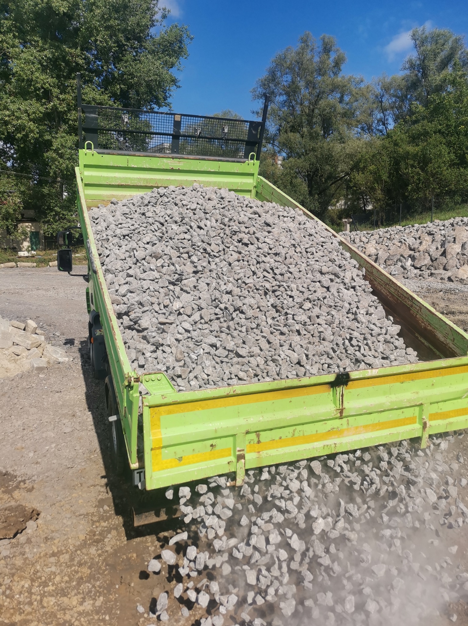 Ciężarówka z otwartą skrzynią ładunkową, wypełniona szarym kruszywem, wysypuje materiał na ziemię, w tle drzewa i budynki.