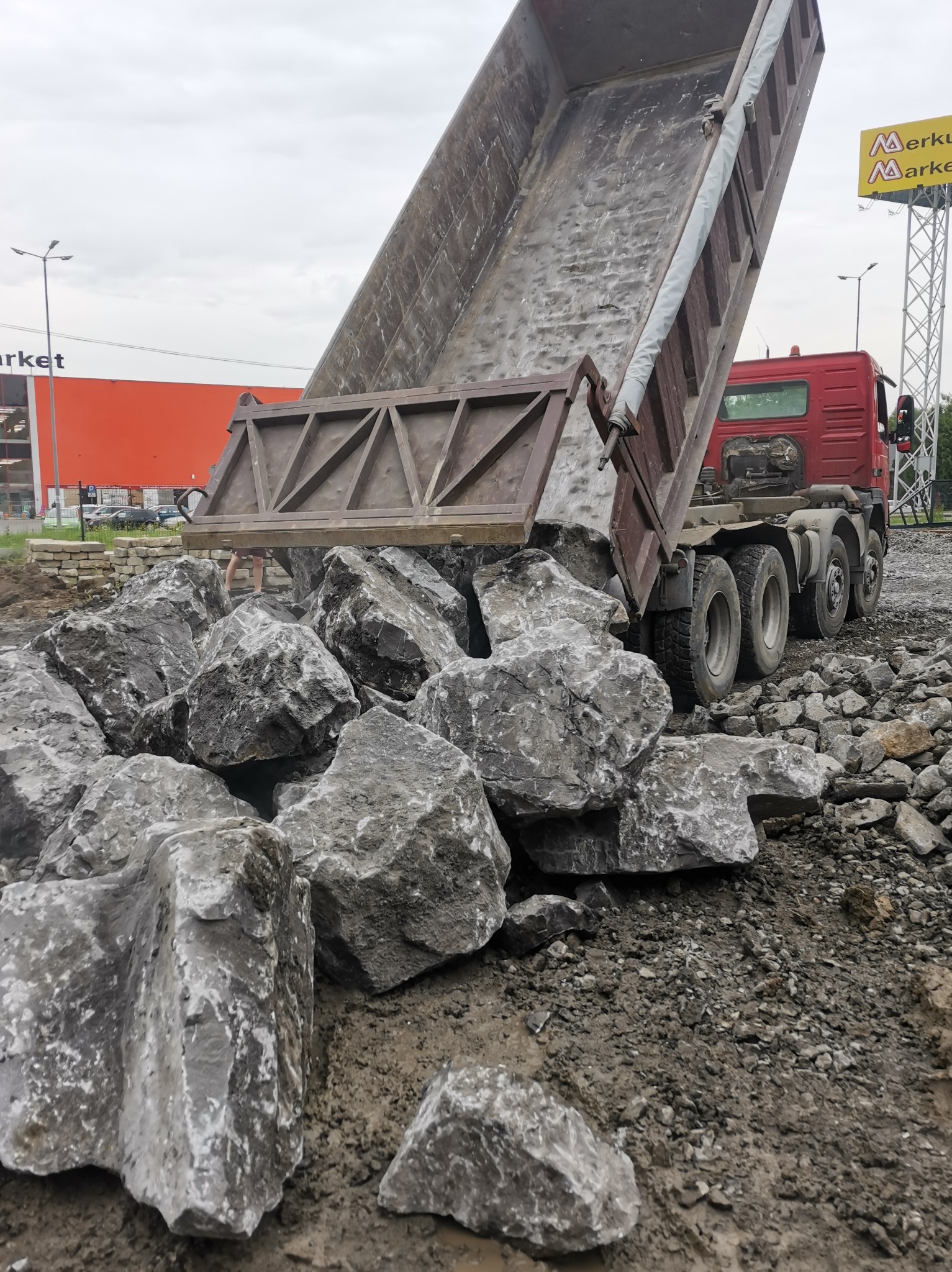 Ciężarówka z uniesioną skrzynią wyładowuje duże, szare kamienie na placu budowy, w tle widoczny budynek sklepu i reklama.