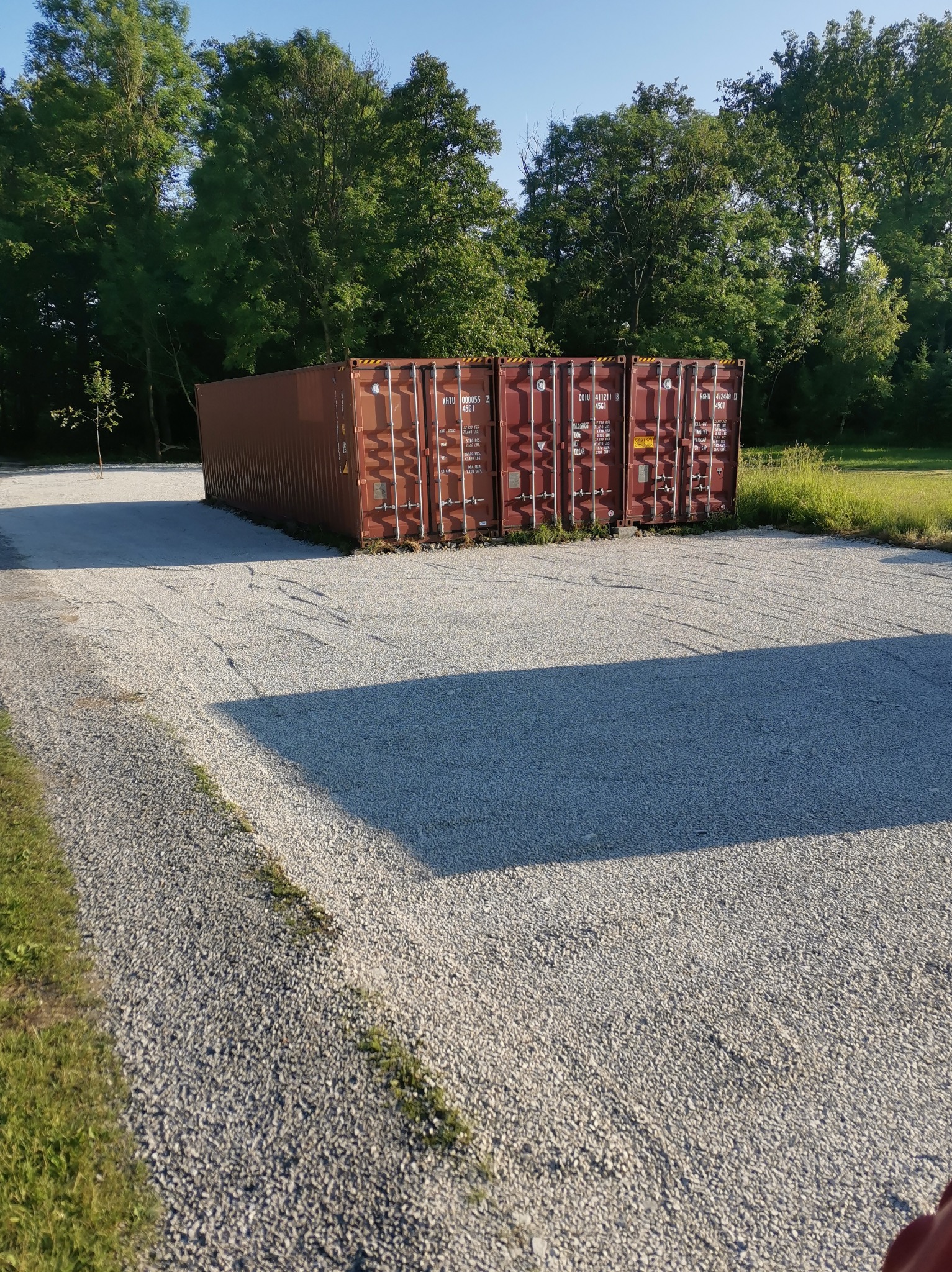 Cztery ustawione obok siebie czerwono-brązowe kontenery morskie na utwardzonym żwirowym placu, otoczone zielenią drzew i trawy.