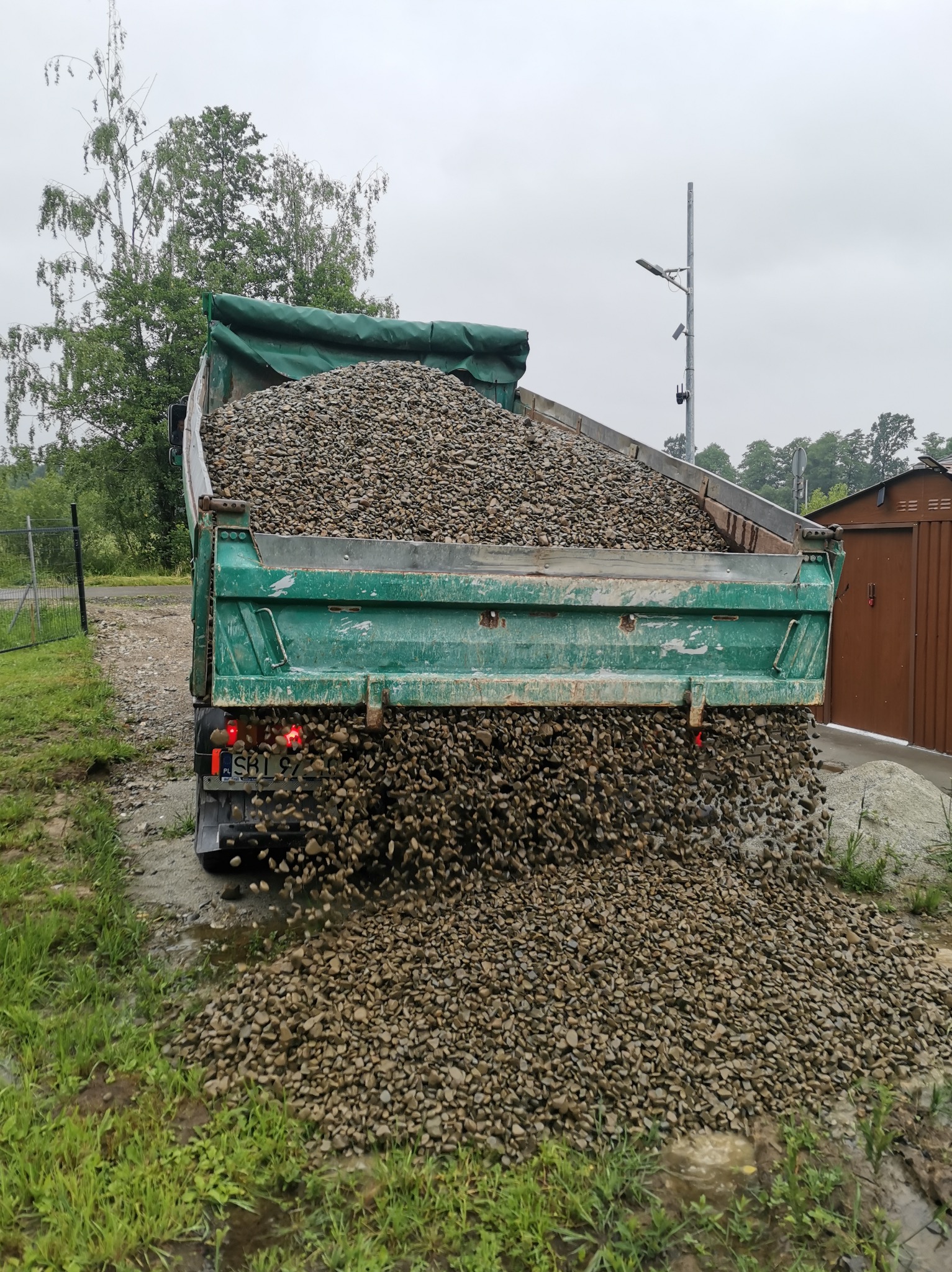 Zielona wywrotka wysypuje żwir na mokry teren z widoczną trawą i kałużami, w tle drzewa i budynek gospodarczy.