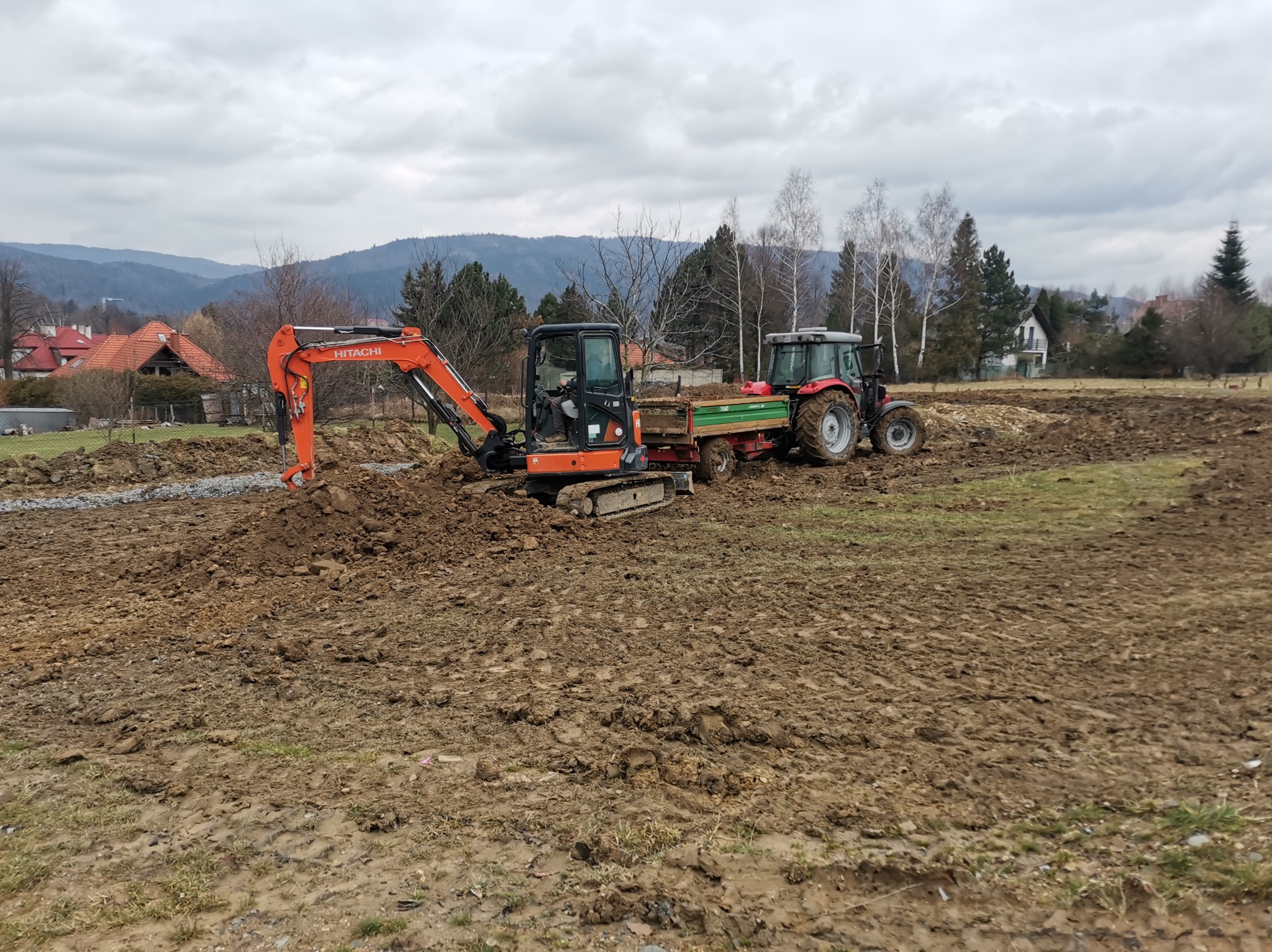 Pomarańczowa mini-koparka Hitachi i czerwony traktor z przyczepą na błotnistym terenie, w tle widoczne drzewa i zabudowania pod pochmurnym niebem.
