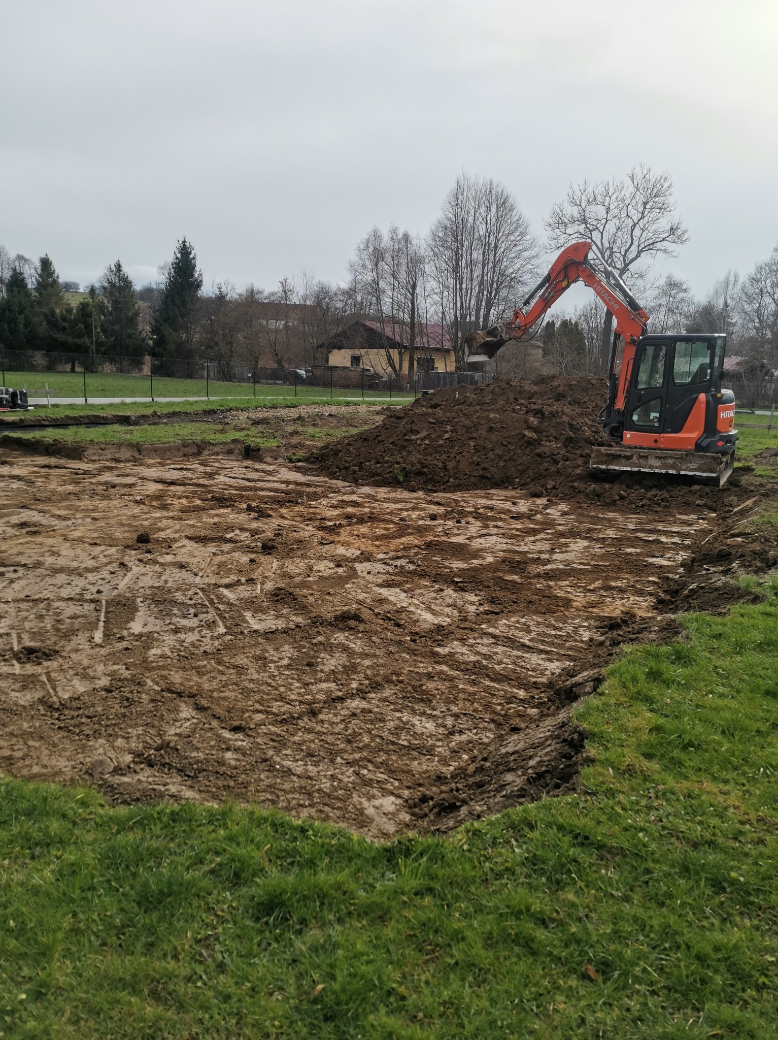 Pomarańczowa mini koparka Hitachi ST51 obok wykopu pod fundamenty, z hałdą ziemi w tle, widok na pole z trawą i zabudowania w oddali, pochmurny dzień.