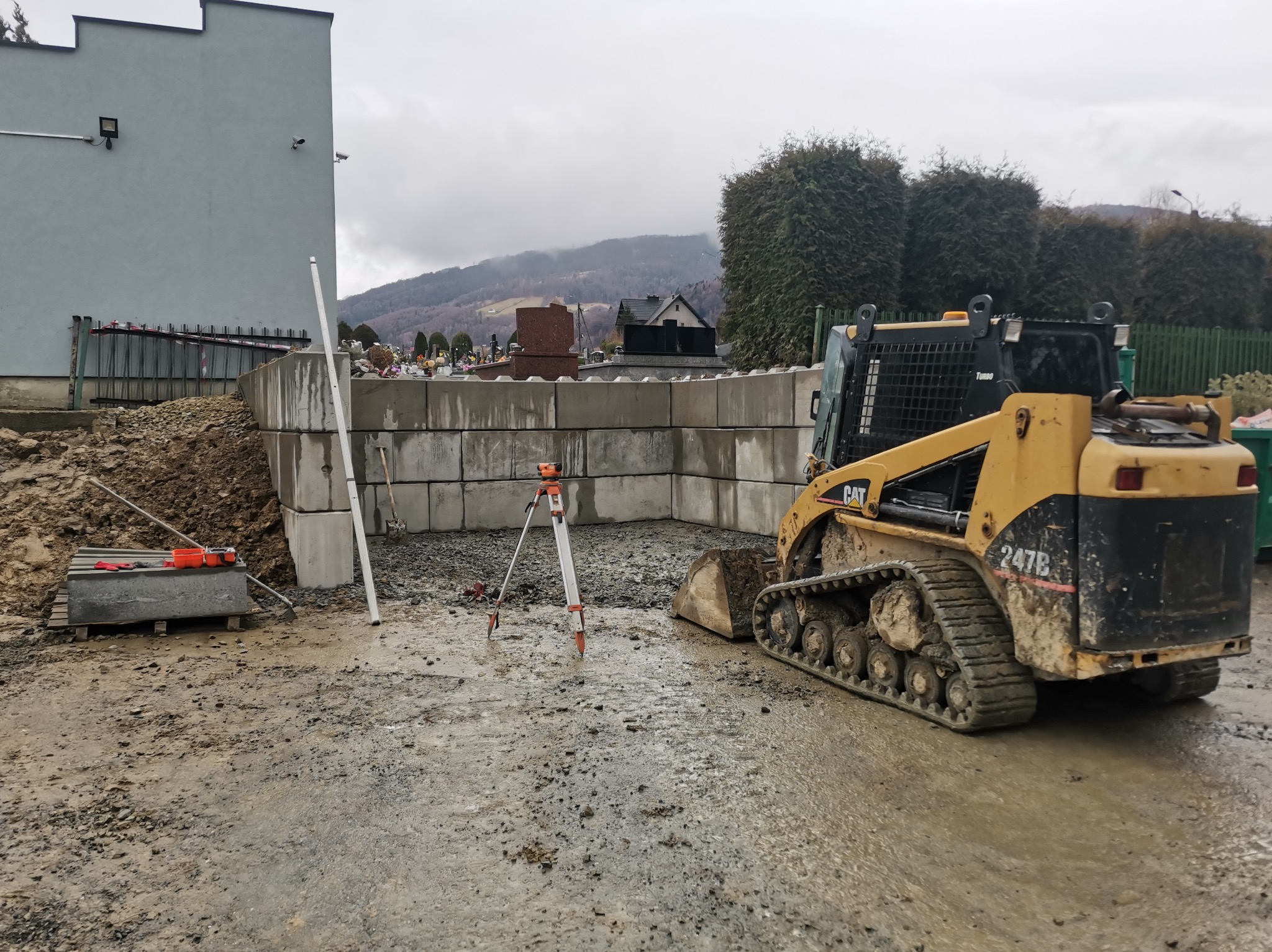 Żółta mini ładowarka gąsienicowa CAT 247B na mokrym placu budowy, w tle mur z betonowych bloków, niwelator optyczny i cmentarz na wzgórzu.