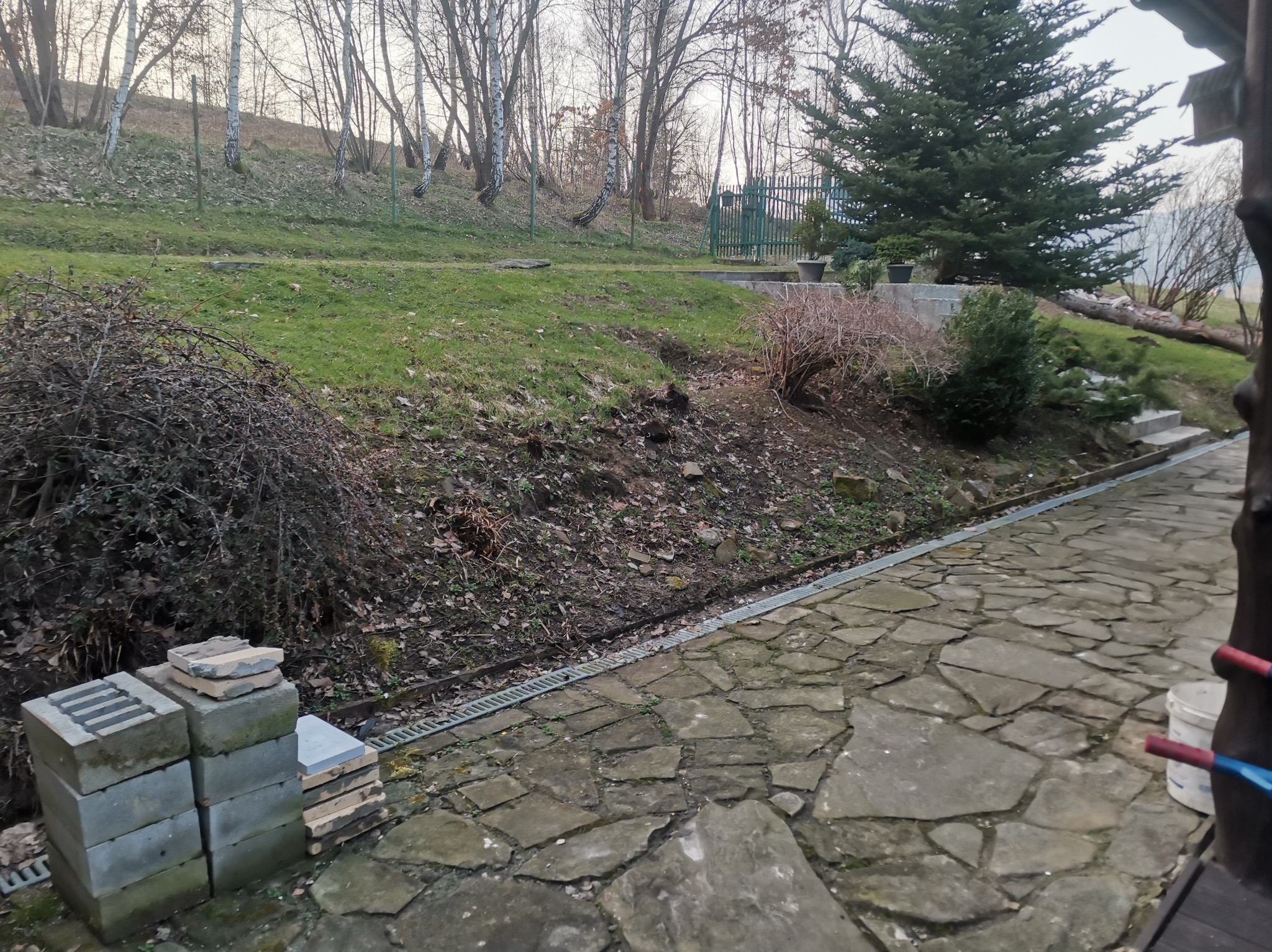 Kamienny chodnik na nierównym terenie z widocznym systemem odwadniającym, ułożone obok siebie bloczki betonowe i kamienne, w tle trawnik z drzewami i krzewami.