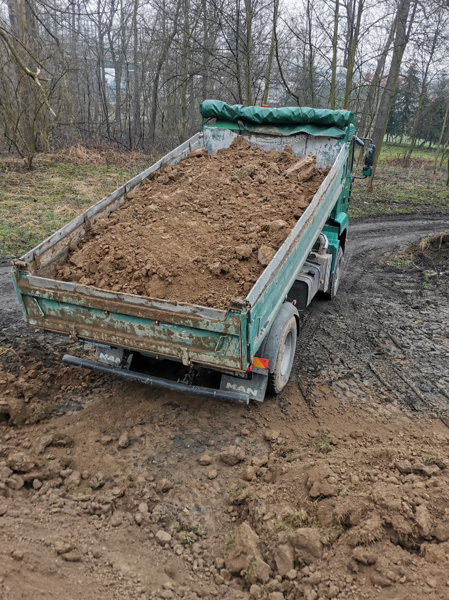 Ciężarówka MAN z zieloną plandeką, wypełniona ziemią, stoi na błotnistej drodze w pobliżu lasu.
