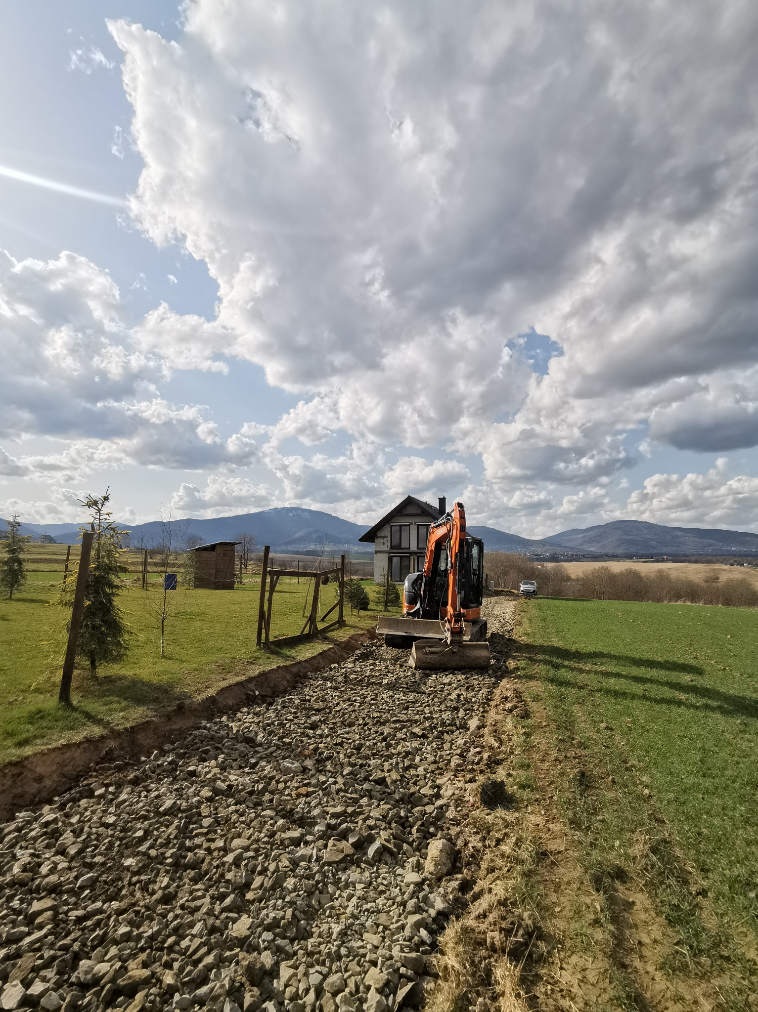 Pomarańczowa minikoparka na żwirowej drodze w trakcie budowy, z domem w tle i malowniczym krajobrazem pagórkowatym pod błękitnym niebem z chmurami.