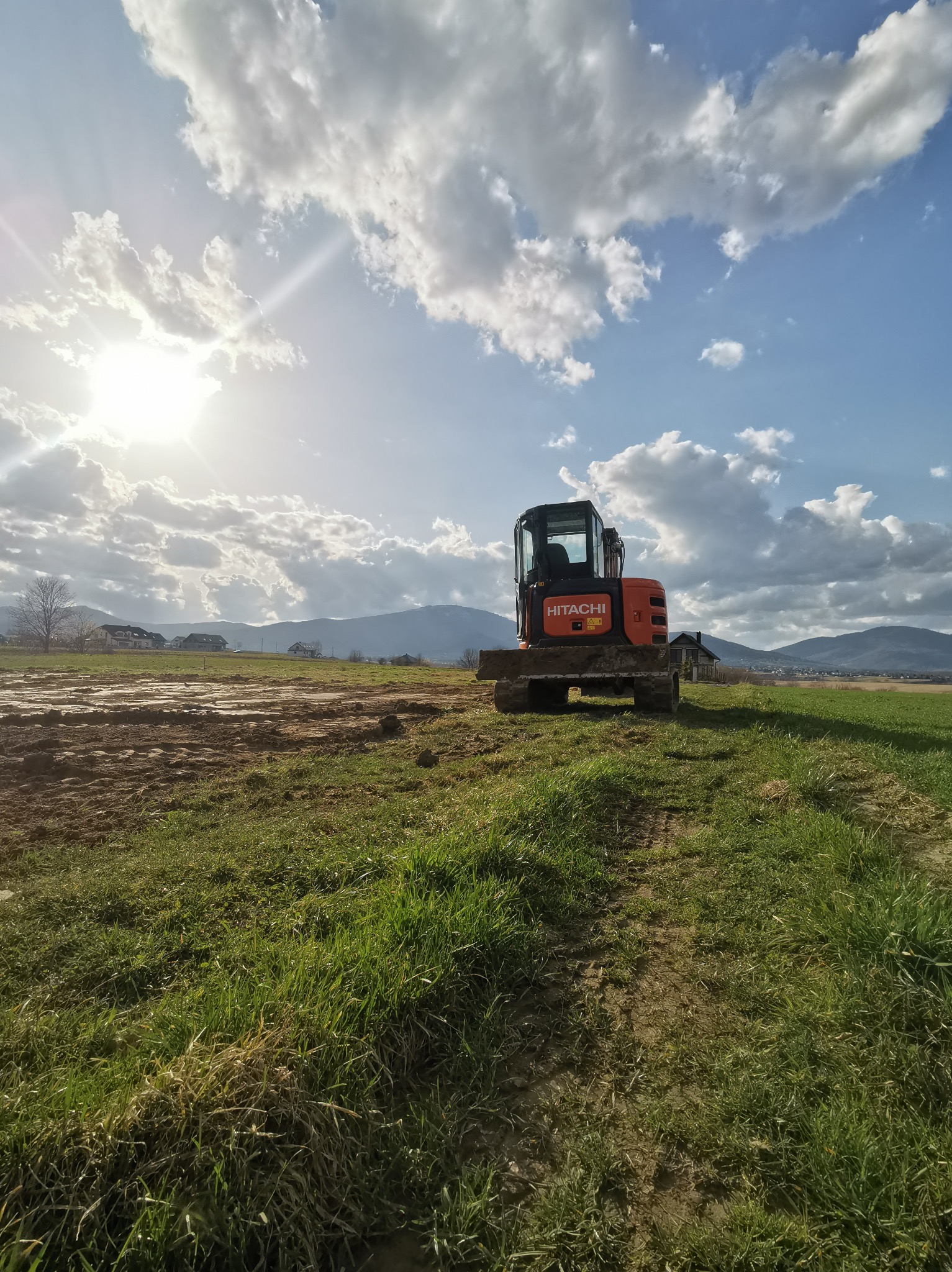 Pomarańczowa mini koparka Hitachi na tle zaoranej ziemi, zielonej trawy, błękitnego nieba z chmurami i gór w oddali.