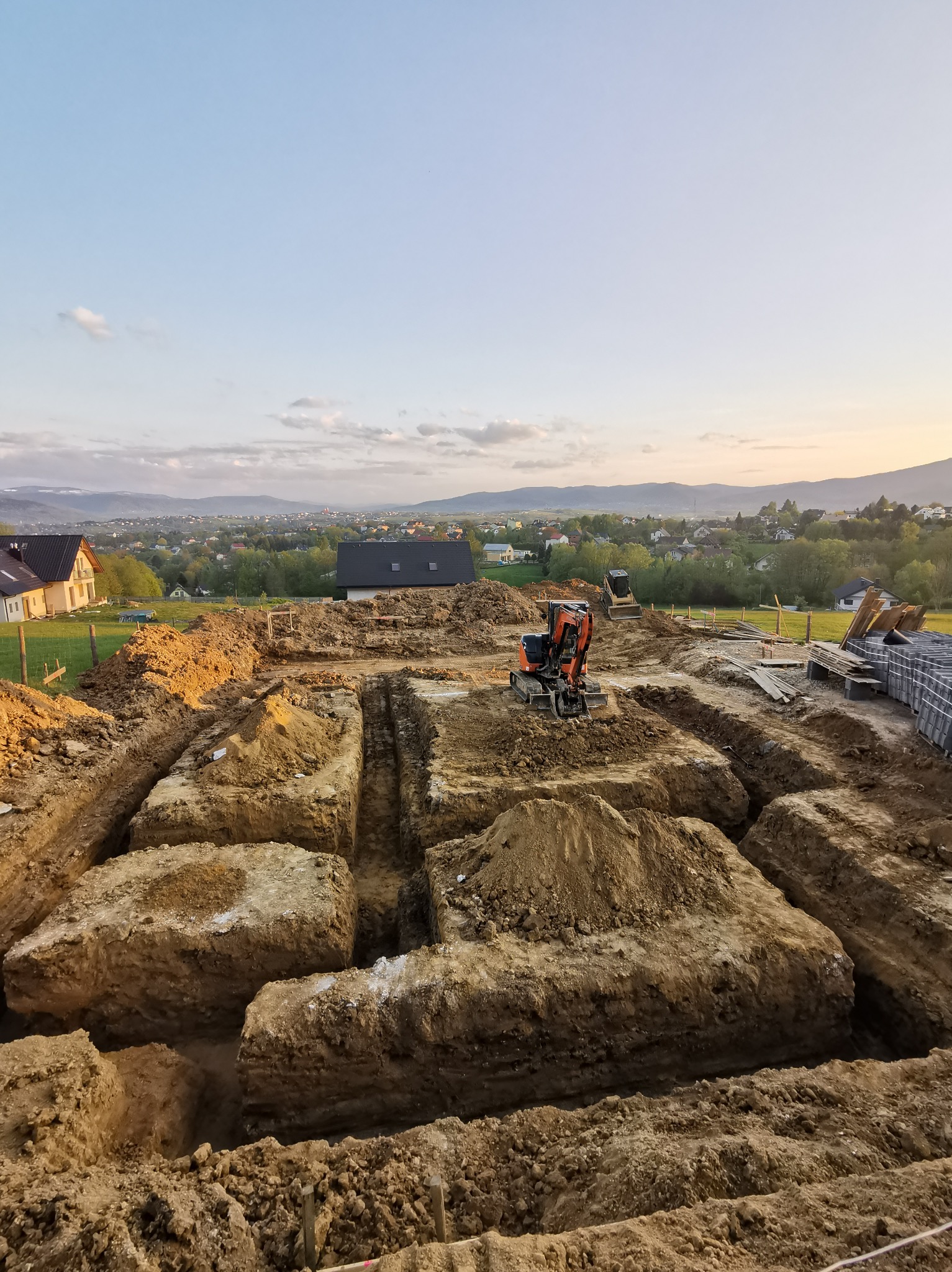 Wykonane wykopy pod fundamenty domu jednorodzinnego z widocznymi dwoma małymi koparkami na tle zabudowy mieszkalnej i wzgórz o zmierzchu.