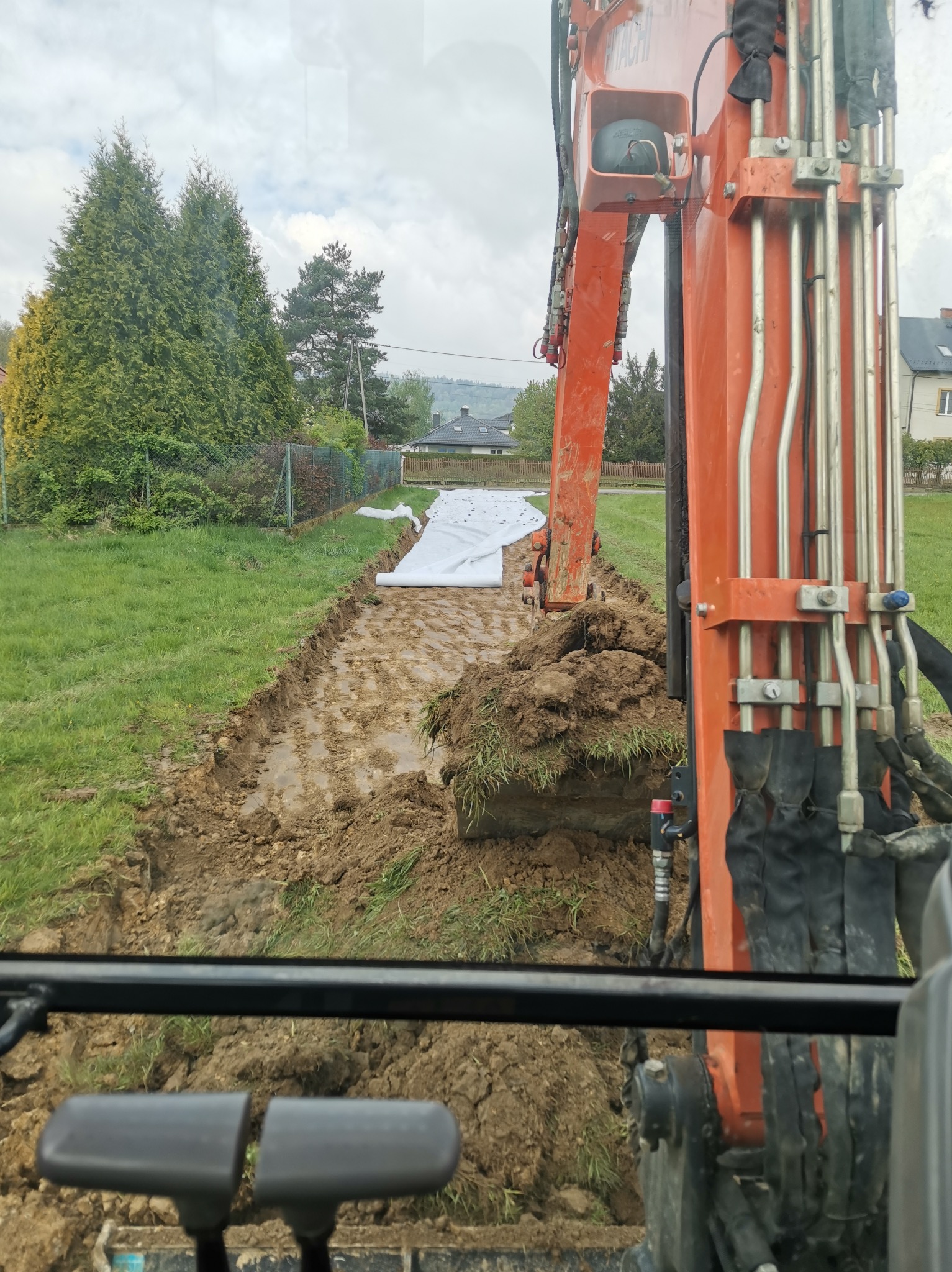Pomarańczowa koparka Hitachi w trakcie wykonywania wykopu w ziemi, widok z kabiny operatora, w tle widoczna biała geowłóknina i zabudowa jednorodzinna.