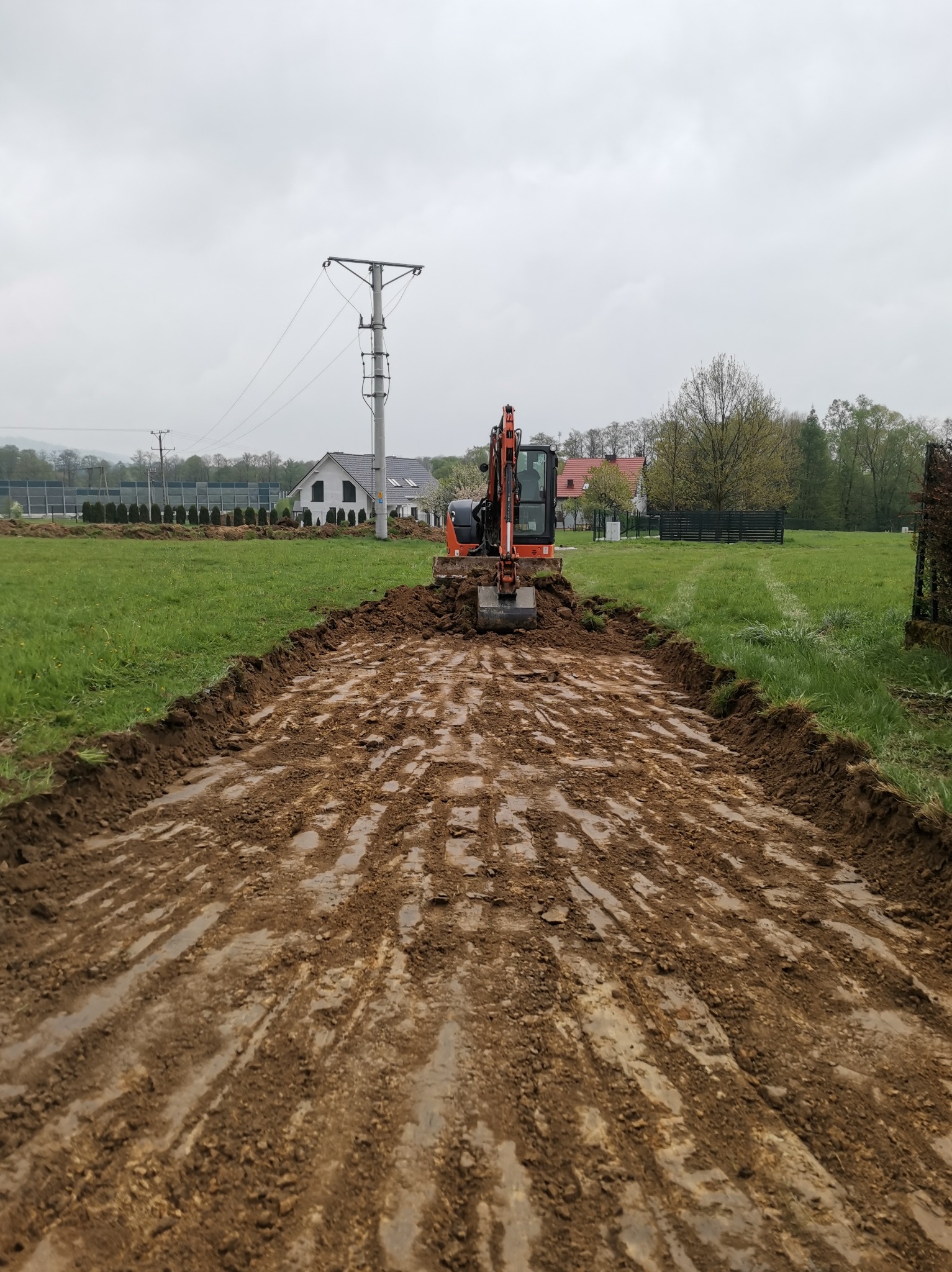 Mini koparka w trakcie prac ziemnych w Rybarzowicach, przygotowanie terenu pod budowę, widoczne ślady opon na ubitej ziemi, w tle dom i linia energetyczna.