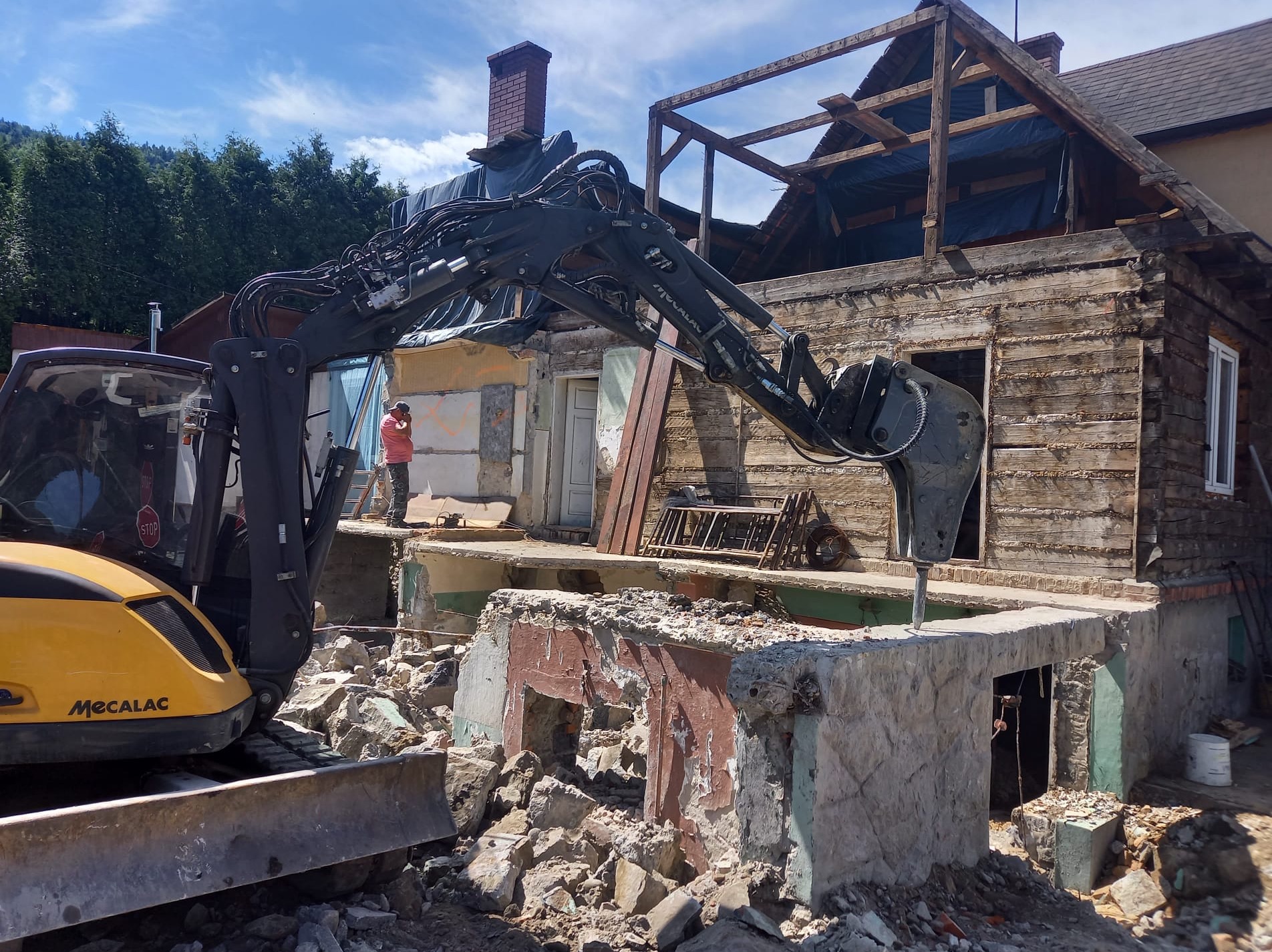 Żółta koparka Mecalac z hydraulicznym młotem pneumatycznym rozbija fundament starego, częściowo rozebranego drewnianego domu z ceglanym kominem. W tle widoczny pracownik w czerwonej koszulce...