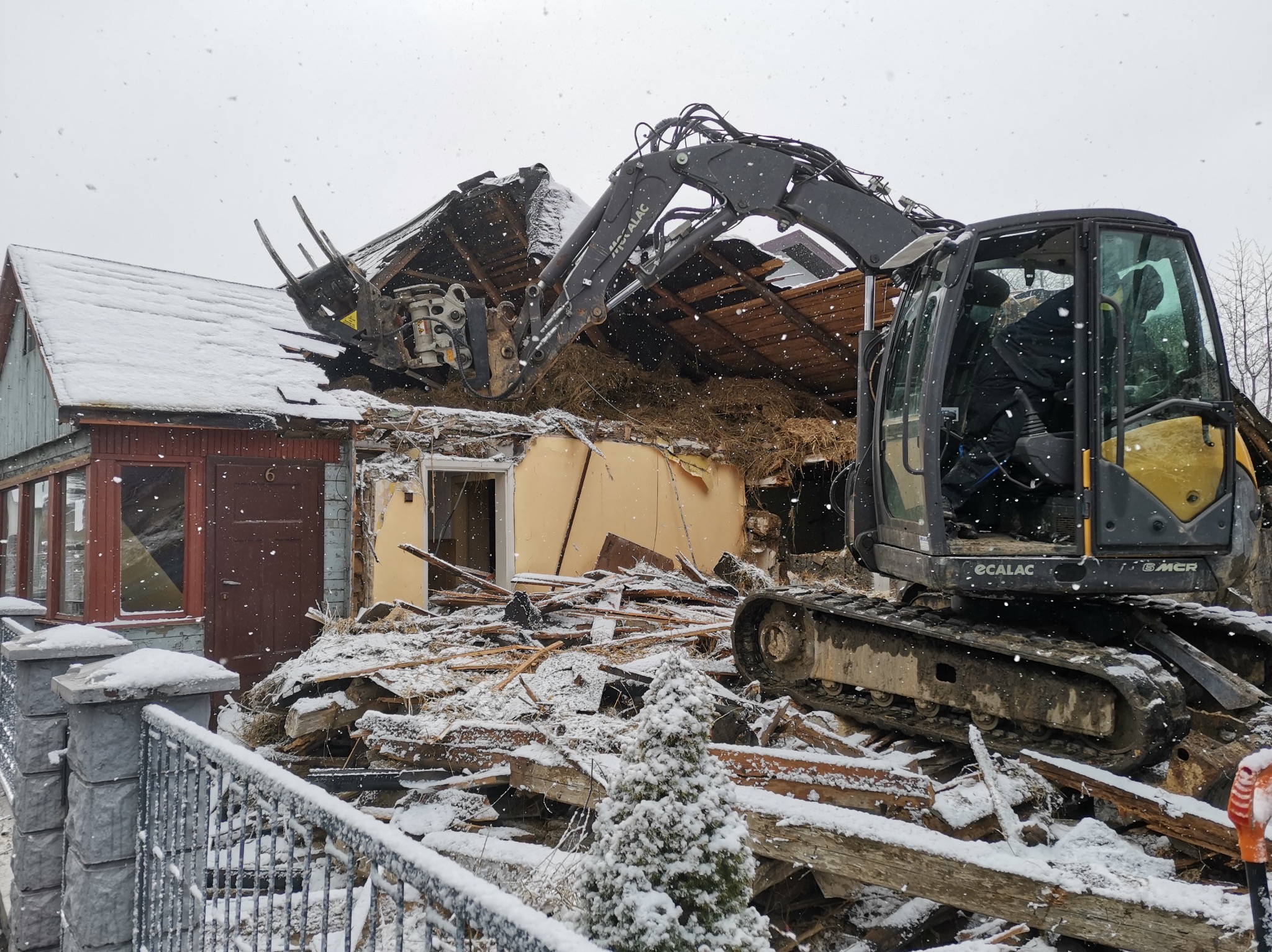 Ciężki sprzęt budowlany rozbiera stary, częściowo zawalony dom w zimowej scenerii, z widocznymi elementami konstrukcyjnymi i gruzem pokrytym śniegiem.