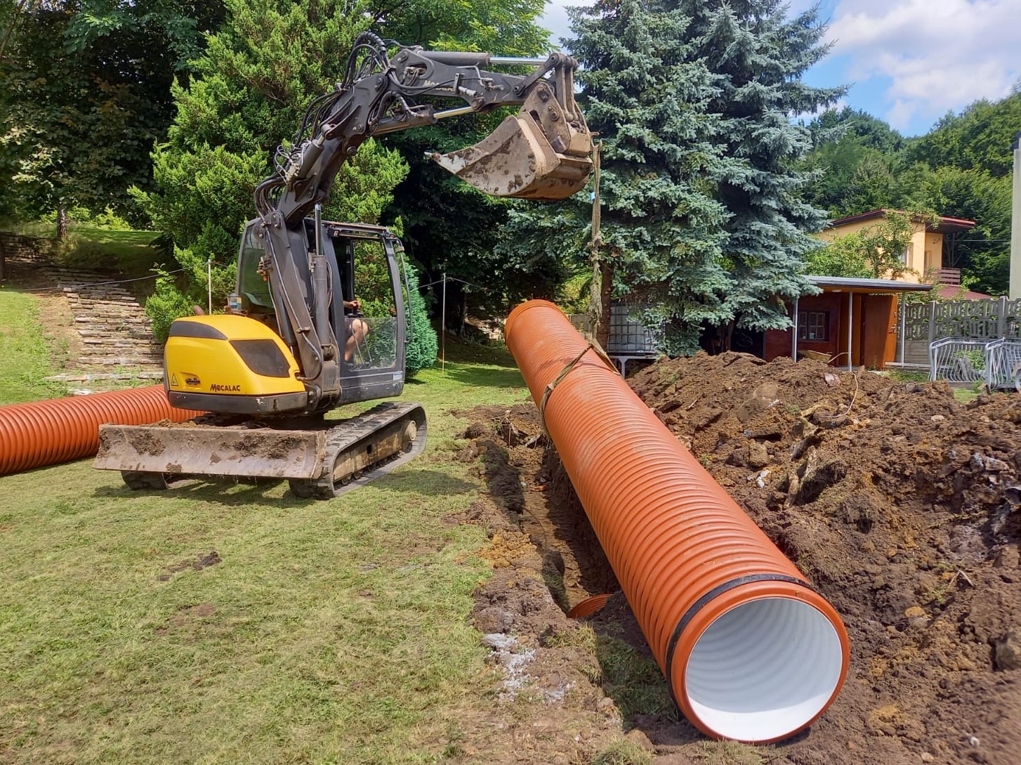 Żółto-czarna koparka układa rurę drenarską w wykopie na trawiastym terenie, w tle drzewa i budynek.