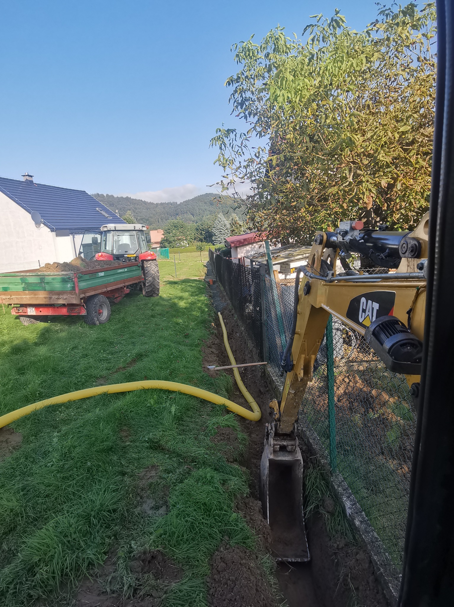 Żółta mini koparka CAT w trakcie wykonywania wąskiego wykopu wzdłuż ogrodzenia z siatki. W tle widoczny traktor z przyczepą wypełnioną ziemią, dom i zalesione wzgórza pod błękitnym niebem.