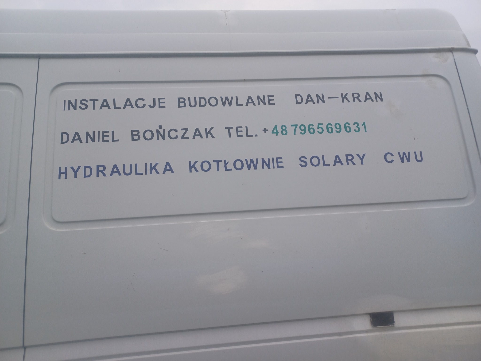 Biały samochód dostawczy z nadrukiem reklamowym: Instalacje Budowlane Dan-Kran, Daniel Bończak, numer telefonu, Hydraulika, Kotłownie, Solary, CWU.