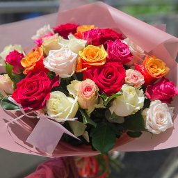 Kwiatowe Atelier Iriska - Kwiaciarnia Wrocław Poczta Kwiatowa - Bukiet mieszanych róż w odcieniach czerwieni, pomarańczy, różu i bieli, owinięty w różowy papier, trzymany w dłoni na tle rozmytego otoczenia.