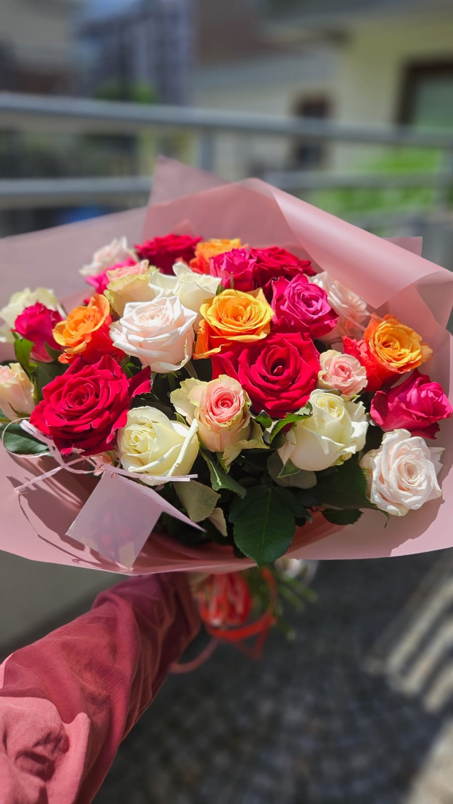 Bukiet mieszanych róż w odcieniach czerwieni, pomarańczy, różu i bieli, owinięty w różowy papier, trzymany w dłoni na tle rozmytego otoczenia.