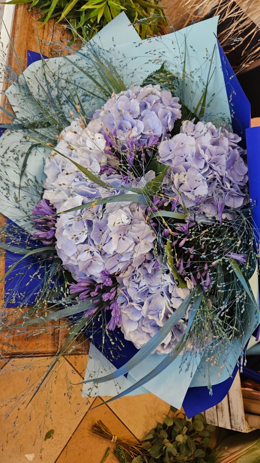 Bukiet z jasnofioletowych hortensji i agapantów, owinięty w niebieski i błękitny papier, widoczny z góry na drewnianym tle.