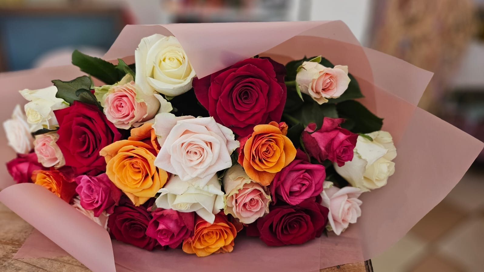 Bukiet mieszanych róż w odcieniach czerwieni, pomarańczy, różu i bieli, zawinięty w delikatny, różowy papier.