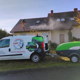 AMKO Marcin Koziarz - Pracownik w kombinezonie czyści dach domu myjką ciśnieniową. W tle biała elewacja i firmowy van z przyczepą. Usługa mycia ciśnieniowego.