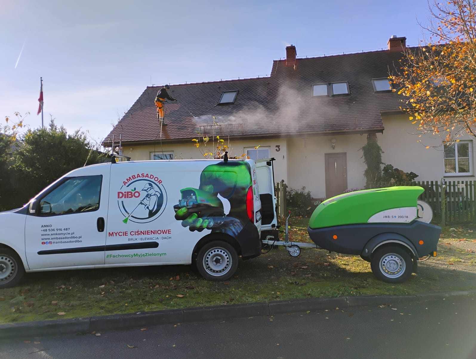 Pracownik w kombinezonie czyści dach domu myjką ciśnieniową. W tle biała elewacja i firmowy van z przyczepą. Usługa mycia ciśnieniowego.