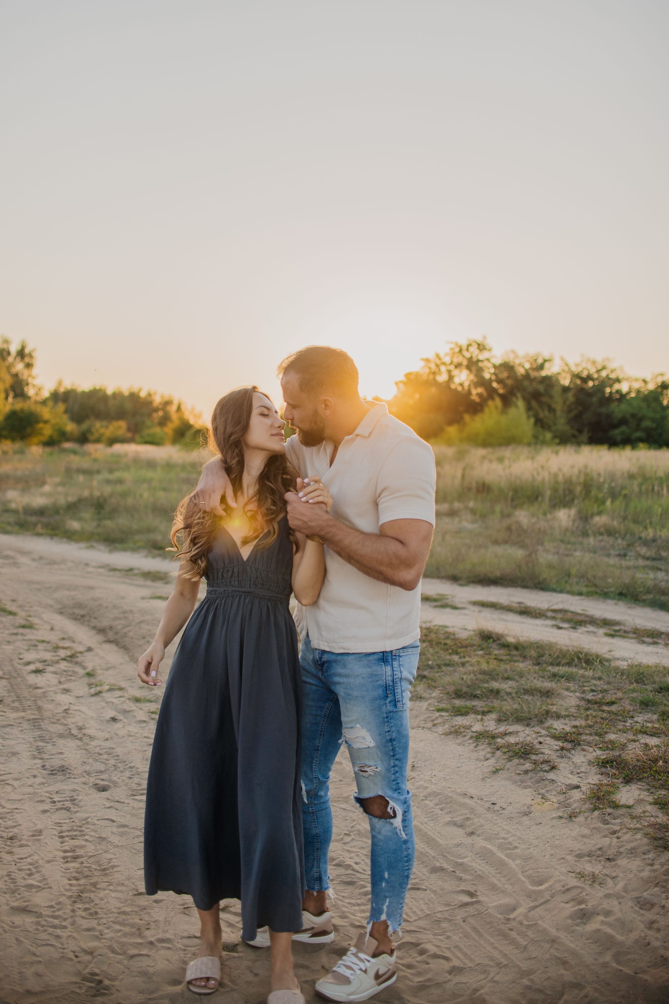 Para zakochanych, mężczyzna w podartych jeansach i kobieta w długiej sukience, trzymają się za ręce na polnej drodze o zachodzie słońca.